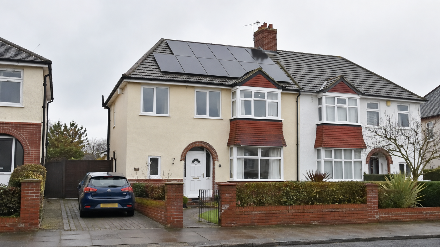 Cream-rendered UK semi-detached house with black solar panels on the front roof, red-brick door arch, dark blue hatchback on the drive